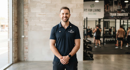 Professional daylight portrait of Tim, male head coach and exercise scientist in navy athletic polo shirt standing against clean industrial neutral wall at Fit For Living gym Geelong, confident warm expression.