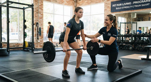A young Australian teenager being coached patiently on clean barbell mechanics by a qualified trainer in navy polo in a sunlit gym facility.